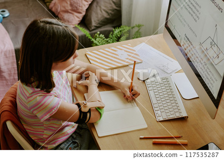 High angle view of teenager with prosthetic arm sitting at desk in her room and writing in copybook during online class High angle view of teenager with prosthetic arm sitting at desk in her room and writing in copybook during online class 117535287