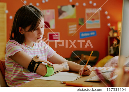 Medium shot of teen girl with artificial arm sitting at desk in her room and doing homework task in copybook 117535304