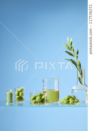 Frontal shot photo against on blue background with olive and yellow oil contained in lab glassware. Olive cosmetics leverage its natural properties to nourish and rejuvenate the skin Frontal shot photo against on blue background with olive and yellow oil contained in lab glassware. Olive cosmetics leverage its natural properties to nourish and rejuvenate the skin 117536251