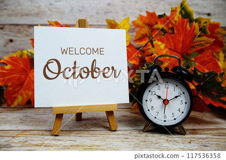 Welcome October text with alarm clock and maple leaf decoration on wooden background Welcome October text with alarm clock and maple leaf decoration on wooden background 117536358