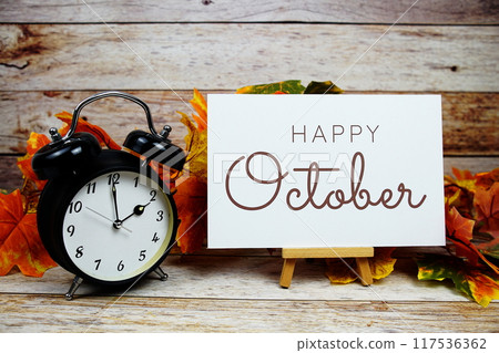 Happy October text with alarm clock and maple leaf decoration on wooden background Happy October text with alarm clock and maple leaf decoration on wooden background 117536362