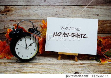 Welcome November text with alarm clock and maple leaf decoration on wooden background 117536363
