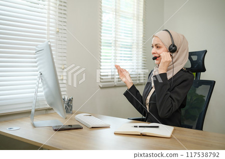 A woman wearing a hijab headset is talking on the phone while sitting at a desk A woman wearing a hijab headset is talking on the phone while sitting at a desk 117538792
