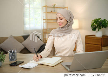 A woman wearing a head scarf is sitting at a desk with a laptop and a notebook 117538793