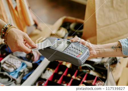 Hand holding payment card while completing transaction through point-of-sale device in retail setting involving jewelry display. Close-up showing customer paying using electronic payment method 117539827