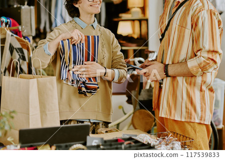 Two young men engaging in conversation while examining cloth hanging in store. Items displayed on table and hanging clothes visible in background, creating retail ambiance 117539833
