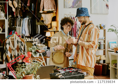 Friends exploring vintage store shelves, admiring various items and collectibles. Clothing racks and eclectic decor create cozy environment, fostering casual, friendly atmosphere Friends exploring vintage store shelves, admiring various items and collectibles. Clothing racks and eclectic decor create cozy environment, fostering casual, friendly atmosphere 117539834