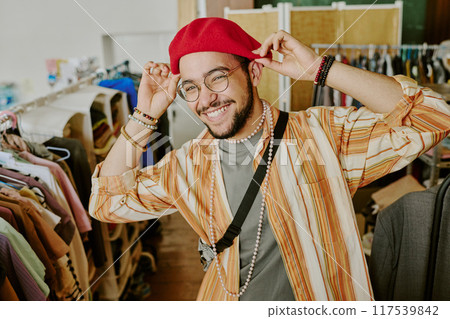 Smiling man wearing red hat and glasses, adjusting hat in clothing store with various apparel on racks surrounding him. Multiracial young man demonstrating vibrant sense of fashion 117539842