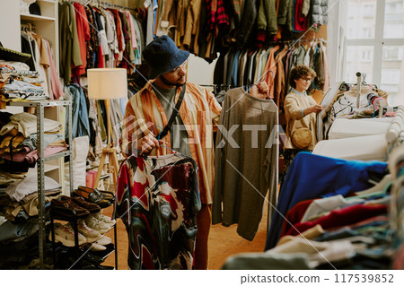 Person browsing racks of clothing in bright thrift store with various garments on display during daylight Peering at different shirts and examining styles and options Person browsing racks of clothing in bright thrift store with various garments on display during daylight Peering at different shirts and examining styles and options 117539852
