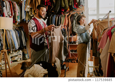 Man and woman browsing clothes and selecting items in vintage clothing store with racks full of colorful outfits surrounding them and lamp lighting the space Man and woman browsing clothes and selecting items in vintage clothing store with racks full of colorful outfits surrounding them and lamp lighting the space 117539885