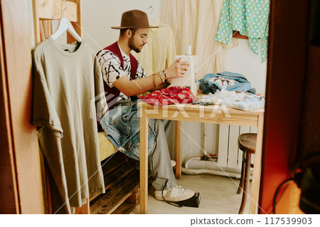 Young adult sitting in small workspace, surrounded by clothing and fabrics, working on new fashion design using creativity and artistic skills Young adult sitting in small workspace, surrounded by clothing and fabrics, working on new fashion design using creativity and artistic skills 117539903