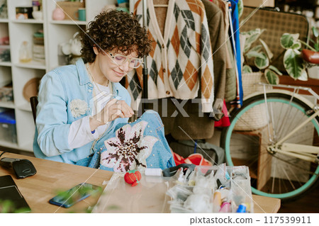 Woman wearing glasses, working on detailed floral graphic embroidery at wooden workspace surrounded by tools, colorful threads, books, and plants adding to cozy atmosphere Woman wearing glasses, working on detailed floral graphic embroidery at wooden workspace surrounded by tools, colorful threads, books, and plants adding to cozy atmosphere 117539911