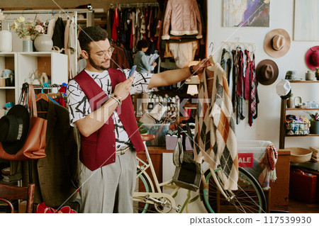 Young man smiling and holding a sweater while browsing clothes in cozy secondhand shop filled with various items and accessories. Multiple people browsing in background Young man smiling and holding a sweater while browsing clothes in cozy secondhand shop filled with various items and accessories. Multiple people browsing in background 117539930