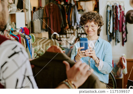 Woman using smartphone to take photo in vintage clothing store. Smiling while focusing on garment held in front, with various clothes and accessories on display around 117539937