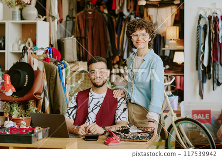 Two smiling people posing in diverse and eclectic vintage clothing store surrounded by various clothes and accessories adding lively mood Two smiling people posing in diverse and eclectic vintage clothing store surrounded by various clothes and accessories adding lively mood 117539942