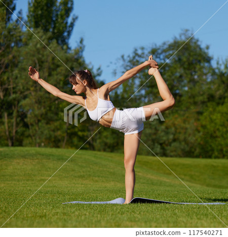 Immersed in yoga routine, beautiful woman works on improving core stability and flexibility outdoor in sunny summer day. 117540271