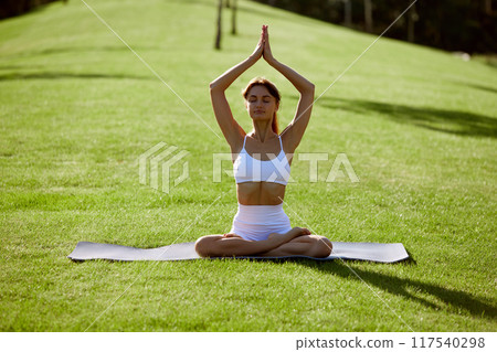 Calm, young woman sitting in lotus, yoga pose on green grass at park, demonstrating wellness. Meditation session outdoor. Calm, young woman sitting in lotus, yoga pose on green grass at park, demonstrating wellness. Meditation session outdoor. 117540298