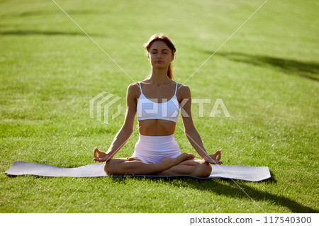 Calm and quiet morning. Calm woman sitting in lotus, yoga pose on green grass at park, demonstrating wellness. Meditation session outdoor. 117540300