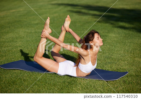 Calm and quiet morning. Flexible, beautiful young woman doing yoga exercises for spine at park bathed in sunbeams. 117540310