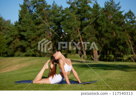 Dynamic photo of young flexible woman doing exercises for back, lying on green grass in sunny morning at park. 117540313