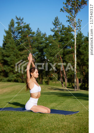 Beautiful, slim woman in comfy activewear sitting on grass in sunny morning at park, doing yoga exercises to fill with energy.. 117540317