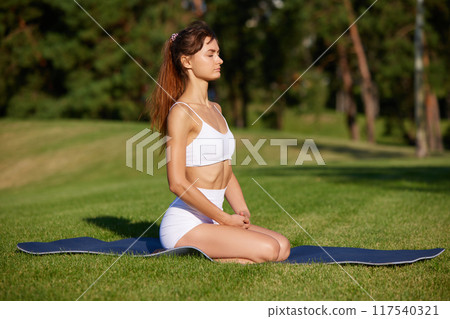 Calm and concentrated on her breathing, young lady sitting with closed eyes on matte on green grass on sunny morning at park. Calm and concentrated on her breathing, young lady sitting with closed eyes on matte on green grass on sunny morning at park. 117540321