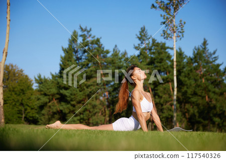 Young fit woman, in white fit wear doing yoga exercises, stretching on green grass at park against blurred background of forest. Young fit woman, in white fit wear doing yoga exercises, stretching on green grass at park against blurred background of forest. 117540326