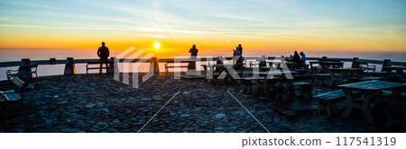 A group of individuals stands at a viewpoint, taking in the breathtaking view of the sunset. Warm hues paint the sky as the sun descends, creating a perfect moment for reflection and connection. 117541319