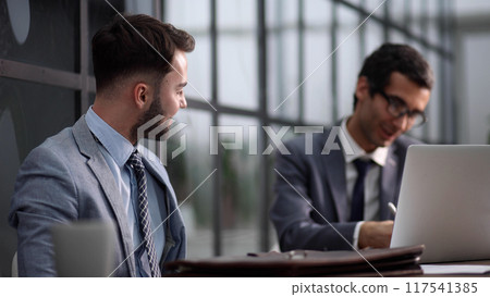 Two serious men in suits sitting at desk with laptop computer and talking. Two serious men in suits sitting at desk with laptop computer and talking. 117541385