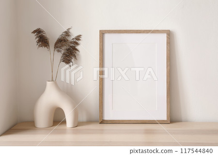 Dry grass, reed flowers bouquet in modern ceramic vase. Empty picture frame, poster mockup on wooden table. White wall background. Minimalistic Scandinavian style, elegant boho interior, living room. Dry grass, reed flowers bouquet in modern ceramic vase. Empty picture frame, poster mockup on wooden table. White wall background. Minimalistic Scandinavian style, elegant boho interior, living room. 117544840