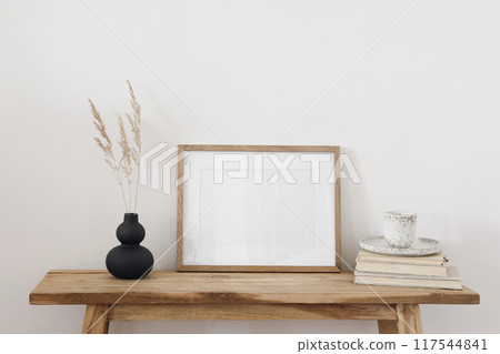 Modern black shaped vase, dry grass flowers on old wooden table, bench. Cup of coffee, books. Horizontal empty picture frame mockup. Art, poster display. Modern interior. White wall background. Modern black shaped vase, dry grass flowers on old wooden table, bench. Cup of coffee, books. Horizontal empty picture frame mockup. Art, poster display. Modern interior. White wall background. 117544841