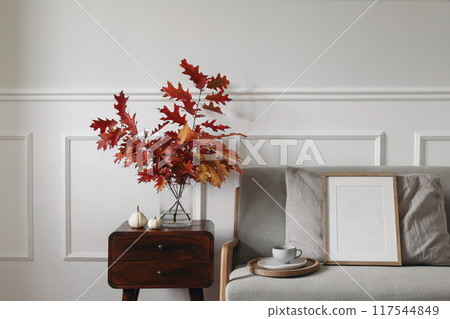 Red oak tree leaves, branches in vase on wooden night table. Linen midcentury sofa with cup of coffee. Blank wooden picture frame mockup. Little white pumpkins. Autumn interior. Thanksgiving 117544849