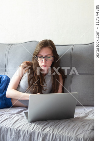 A young woman with glasses lies on the sofa and communicates via video conference via a laptop. A young woman with glasses lies on the sofa and communicates via video conference via a laptop. 117544889