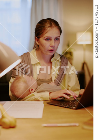 Woman focused on laptop while holding baby in arms, balancing work and childcare in home office environment. Soft lighting from lamp accents cozy workspace with papers spread Woman focused on laptop while holding baby in arms, balancing work and childcare in home office environment. Soft lighting from lamp accents cozy workspace with papers spread 117545313