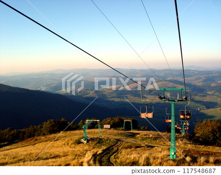 Cable car way against the backdrop of highlands and blue sky. Green cable rack and chair lift ropes Cable car way against the backdrop of highlands and blue sky. Green cable rack and chair lift ropes 117548687