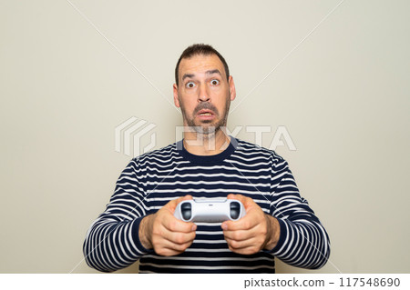 Hispanic man with beard about 40s playing video games with gamepad over isolated beige background scared and surprised, afraid and excited with fear expression. Hispanic man with beard about 40s playing video games with gamepad over isolated beige background scared and surprised, afraid and excited with fear expression. 117548690