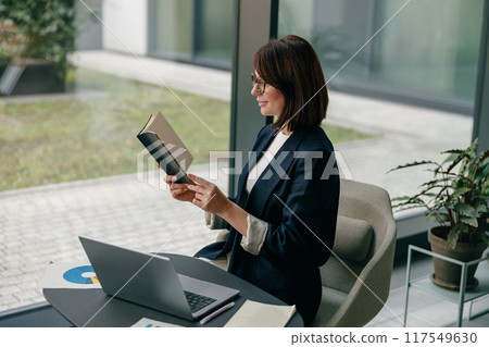A focused businesswoman is reading a book in a modern office surrounded by a laptop and a notebook 117549630
