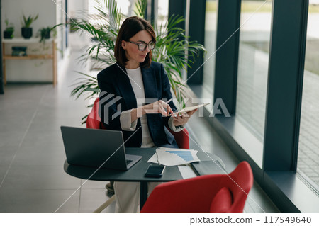 A businesswoman uses a tablet and laptop in her office, focusing on work tasks with dedication 117549640