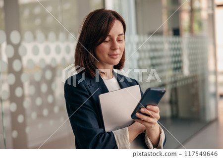 Confident businesswoman is focused on using her smartphone in a modern office setting Confident businesswoman is focused on using her smartphone in a modern office setting 117549646