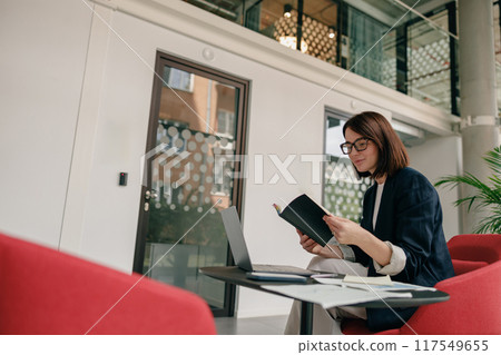 In a contemporary office, a female professional is focused on her work using a laptop and notebook In a contemporary office, a female professional is focused on her work using a laptop and notebook 117549655