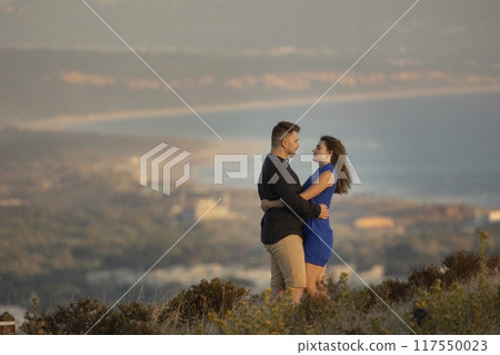 Young couple embracing on a hilltop overlooking a city at sunset 117550023