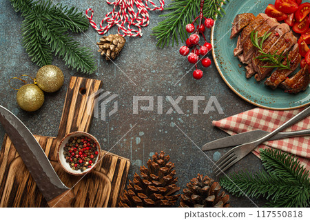 Festive Christmas dinner boarder background with space for text with roasted duck breast fillet with golden crispy skin and tomato salad, Xmas decorations with fir tree, cones, red berries. 117550818