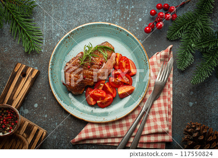 Festive Christmas dinner with roasted duck breast fillet with golden crispy skin and tomato salad, Xmas decorations on table with fir tree, cones, red berries on blue rustic concrete background. Festive Christmas dinner with roasted duck breast fillet with golden crispy skin and tomato salad, Xmas decorations on table with fir tree, cones, red berries on blue rustic concrete background. 117550824