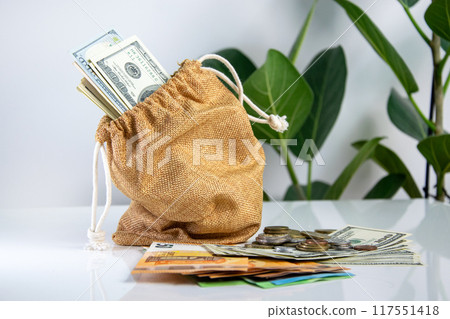 A burlap bag filled with US dollar bills and coins sits on a table with a green plant in the background 117551418