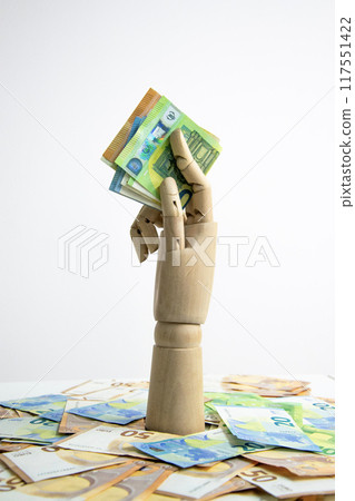 A wooden hand holds several Euro banknotes over a pile of Euro bills 117551422