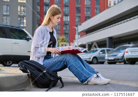 Young college student, girl with backpack reading textbook, outdoors Young college student, girl with backpack reading textbook, outdoors 117551687
