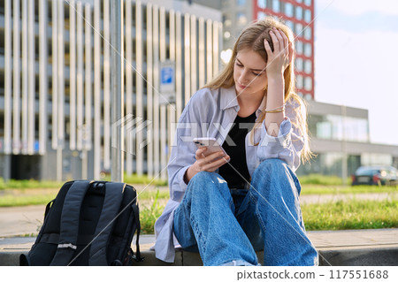 Upset sad unhappy young female using smartphone outdoor, urban style 117551688