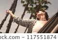 Young woman performs dance movements leaning out of car window traveling along road with palm trees 117551874