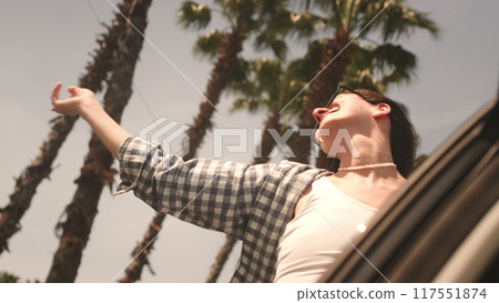 Young woman performs dance movements leaning out of car window traveling along road with palm trees Young woman performs dance movements leaning out of car window traveling along road with palm trees 117551874