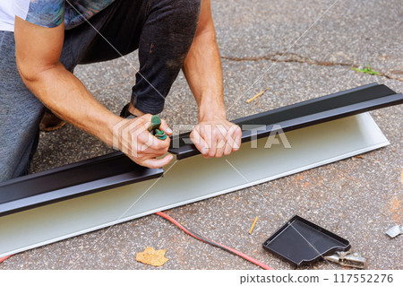 Worker cuts aluminum rain gutters to required size after bending them using machine tool 117552276